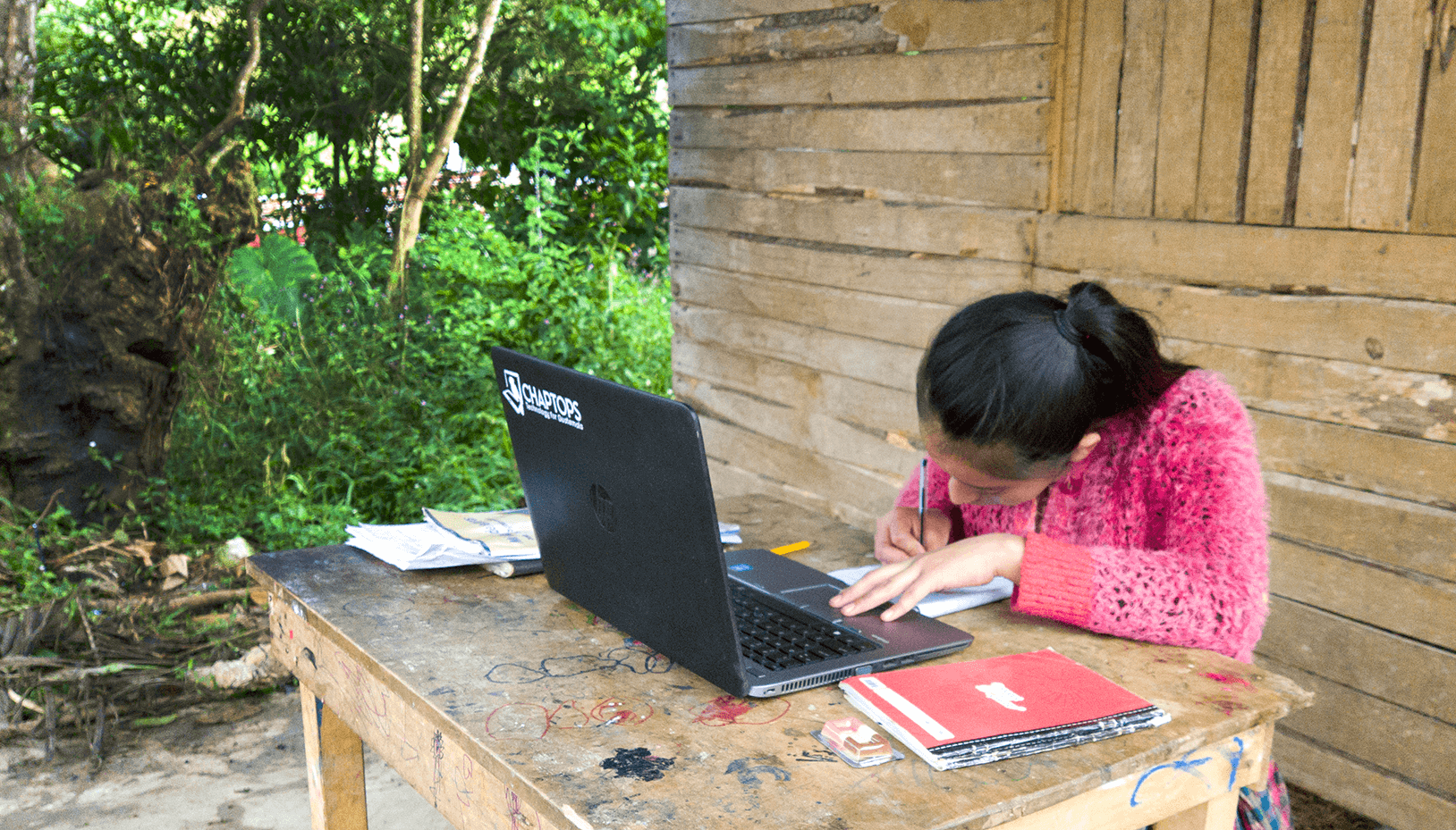 A girl studying with a Chaptops computer at a rough-hewn table in front of a rough-hewn cabin in Alta Verapaz, Guatemala.